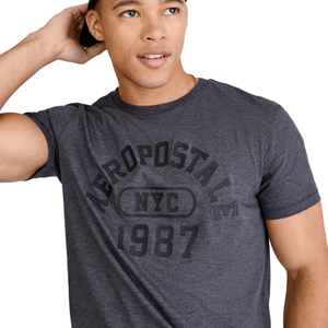 A man wearing a dark gray T-shirt with \“Aéropostale NYC 1987\“ printed in black. He's holding a black cap on his head.