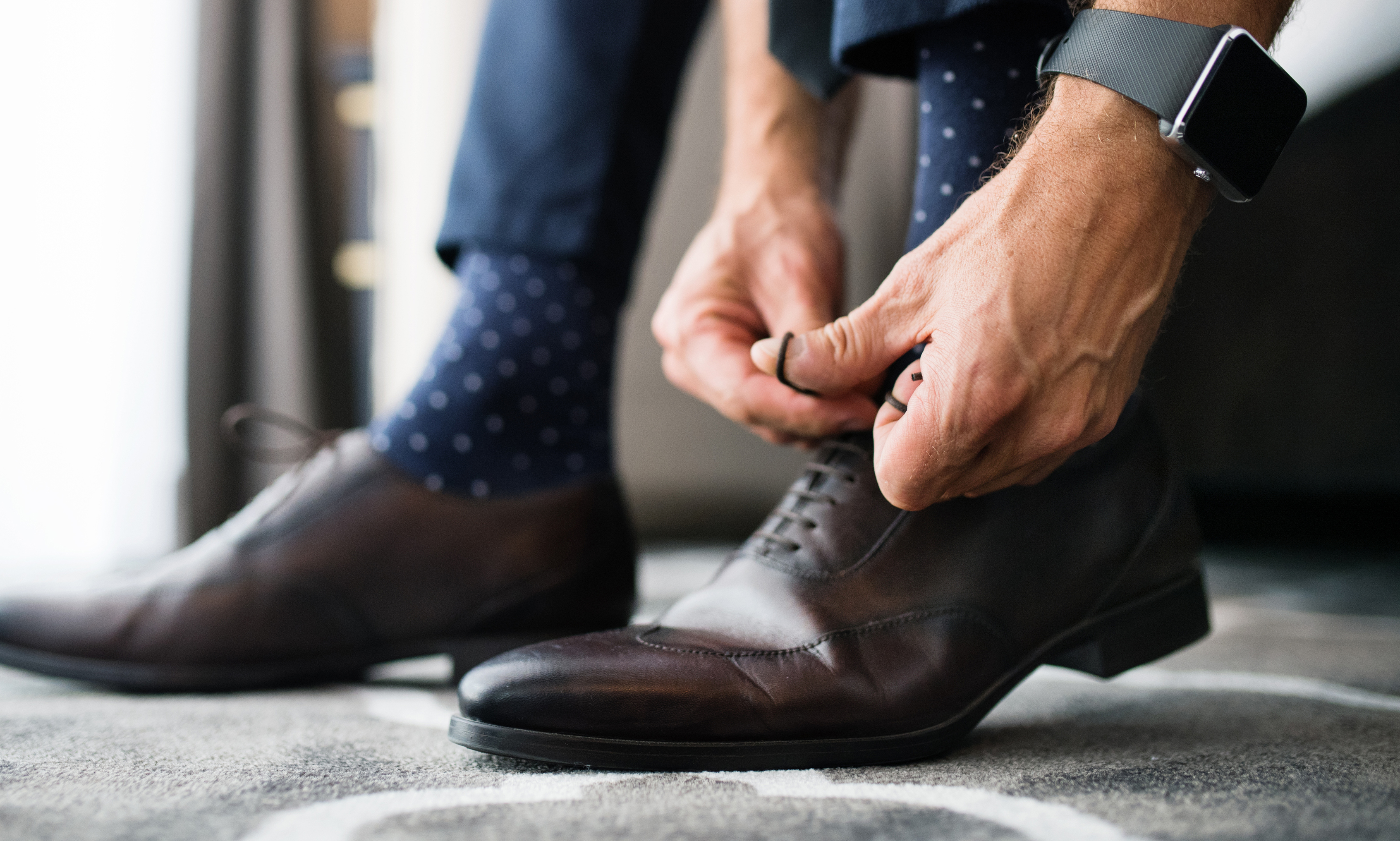 A person tying laces on brown leather dress shoes, wearing blue socks and a smart watch.