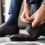 A person tying laces on brown leather dress shoes, wearing blue socks and a smart watch.
