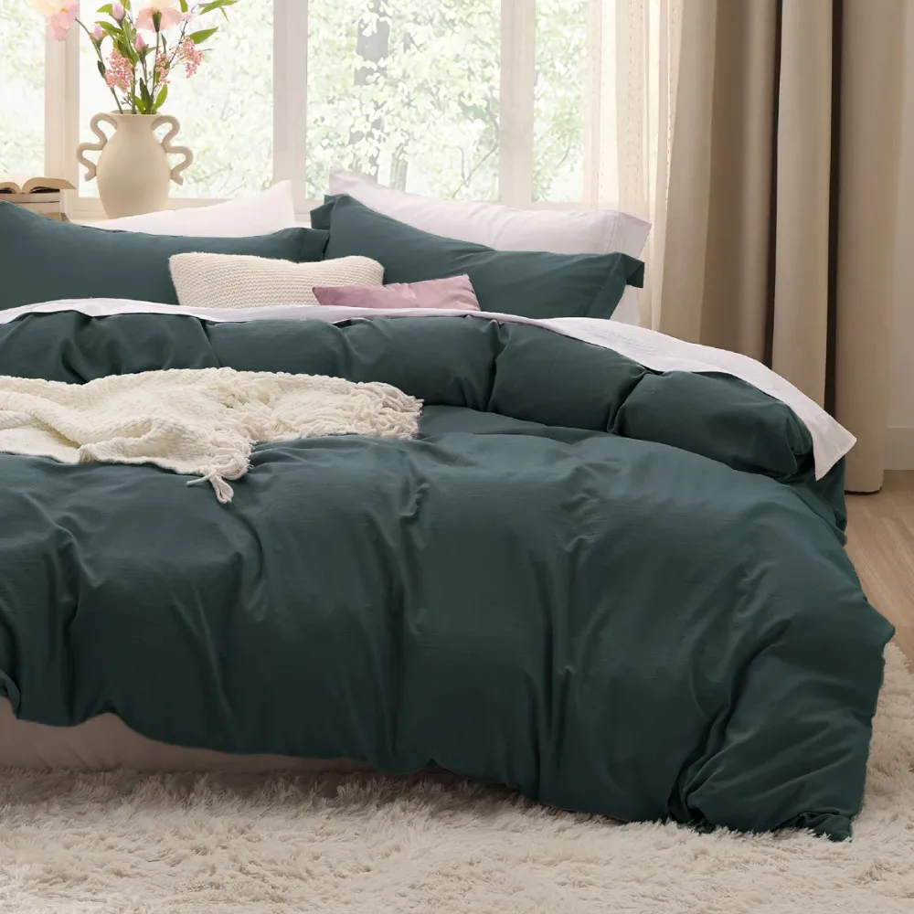 The photo shows a bed with a dark green duvet cover set, featuring multiple pillows and a white textured throw blanket. The room has a cozy and serene ambiance with natural light and decorative flowers in the background.