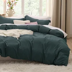 The photo shows a bed with a dark green duvet cover set, featuring multiple pillows and a white textured throw blanket. The room has a cozy and serene ambiance with natural light and decorative flowers in the background.