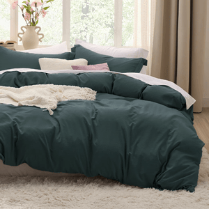 The photo shows a bed with a dark green duvet cover set, featuring multiple pillows and a white textured throw blanket. The room has a cozy and serene ambiance with natural light and decorative flowers in the background.