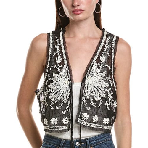A woman is wearing a sleeveless, black mesh vest with intricate white embroidery over a white top and jeans.