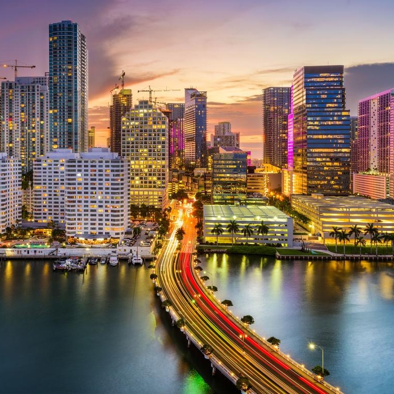 A vibrant cityscape with illuminated skyscrapers reflecting on water at sunset, connected by a bridge with light trails of vehicles.