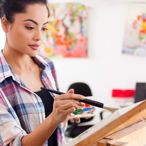 A person is painting on a canvas, holding a palette with various paint colors. They are using a paintbrush and are in a room with colorful abstract paintings in the background.