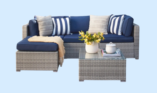 Outdoor wicker patio set featuring a gray sectional sofa with blue cushions, striped and textured pillows, a knitted throw, and a glass-top coffee table adorned with yellow flowers and a teapot.