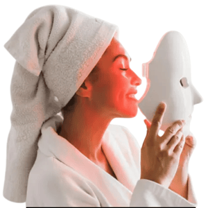 A person with a towel wrapped around their head is wearing a white robe and holding a white LED light therapy mask emitting red light near their face.