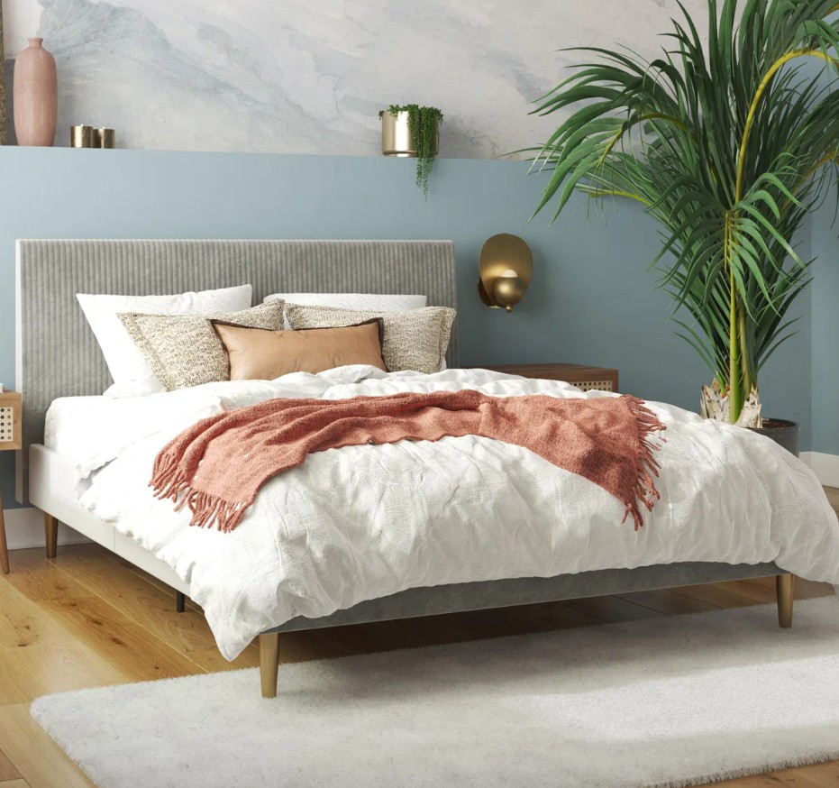 A modern bedroom features a bed with a gray upholstered headboard, white and patterned pillows, a blush throw, a light wooden floor, a white rug, and a large potted plant.