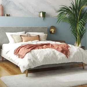A modern bedroom features a bed with a gray upholstered headboard, white and patterned pillows, a blush throw, a light wooden floor, a white rug, and a large potted plant.