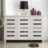 A white finished shoe cabinet with slatted doors, capable of holding up to 16 pairs of shoes, is complemented by a black lamp and decorative bottles on top. Shoes are partially visible underneath.