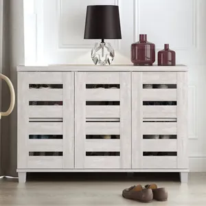 A white finished shoe cabinet with slatted doors, capable of holding up to 16 pairs of shoes, is complemented by a black lamp and decorative bottles on top. Shoes are partially visible underneath.