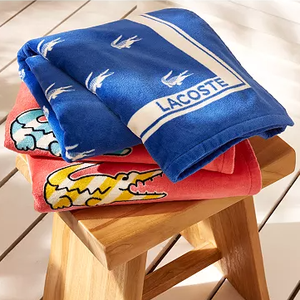 Blue and red towels with Lacoste branding, featuring crocodile and bird designs, are neatly folded on a wooden stool.
