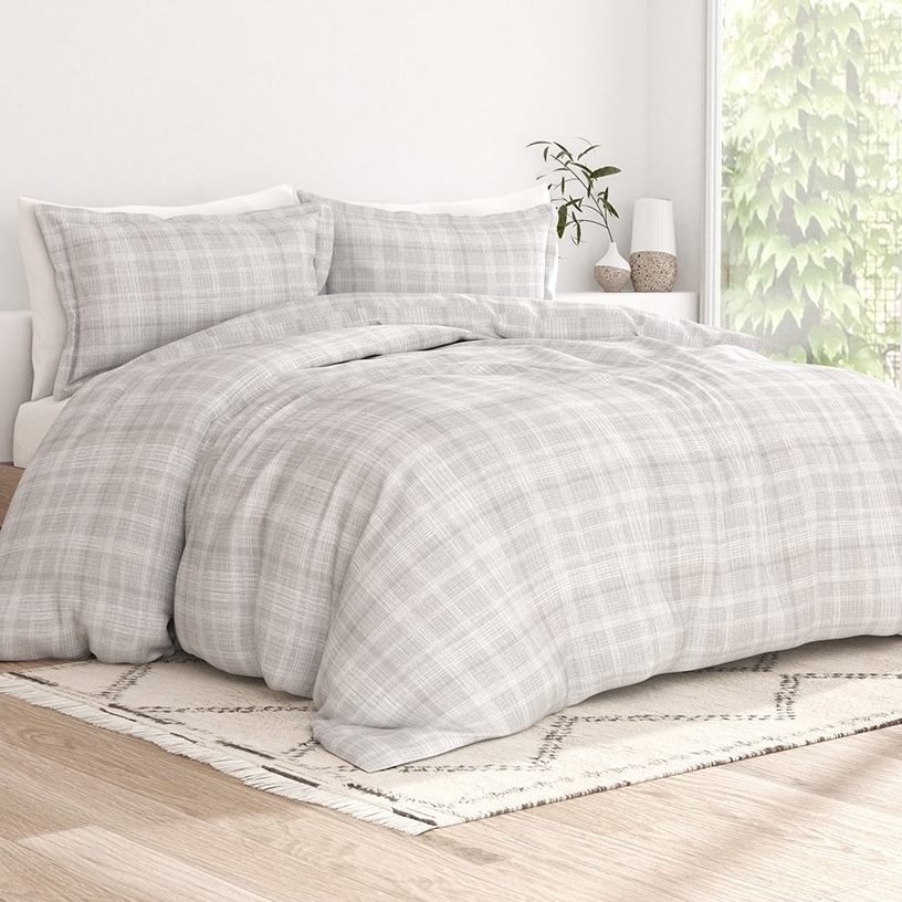 A bedroom setup featuring a bed with a grey plaid duvet cover and matching pillows, a patterned rug on a wooden floor, and a bedside table with a plant and decorative items.