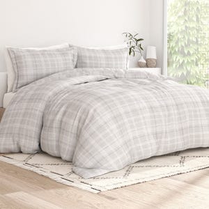 A bedroom setup featuring a bed with a grey plaid duvet cover and matching pillows, a patterned rug on a wooden floor, and a bedside table with a plant and decorative items.