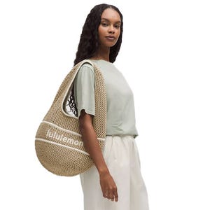 A beige cotton crochet shoulder bag with \“lululemon\“ text, featuring a large curved shape and dual handles, worn on a person's shoulder. Ideal for casual use with ample space for essentials.