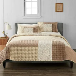A neatly made bed features a patchwork quilt and matching pillows in warm, neutral tones with a side table, lamp, and wall art as decor.