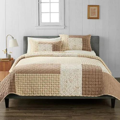 A neatly made bed features a patchwork quilt and matching pillows in warm, neutral tones with a side table, lamp, and wall art as decor.
