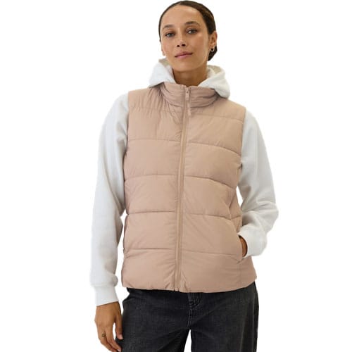 A woman is wearing a light pink, sleeveless ColdControl Puffer Vest from Gap Factory, paired with a white hoodie and dark jeans. The vest features a quilted design with horizontal seams and a front zipper, offering a casual, layered look.