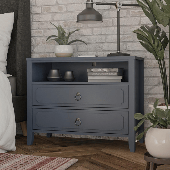 A blue nightstand features two drawers with ring handles, an open shelf holding small decorative bowls and books, a table lamp, and a potted plant. It's placed against a brick wall, with a plant beside it and a patterned rug on a wooden floor.