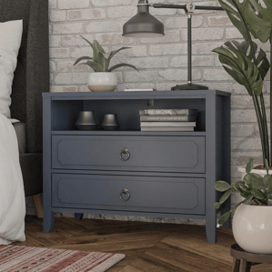 A blue nightstand features two drawers with ring handles, an open shelf holding small decorative bowls and books, a table lamp, and a potted plant. It's placed against a brick wall, with a plant beside it and a patterned rug on a wooden floor.