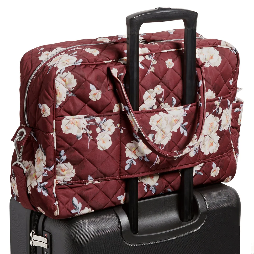 A maroon quilted floral-patterned bag with a handle is attached to the extended handle of a black suitcase. The bag features two carry handles and a zip closure.
