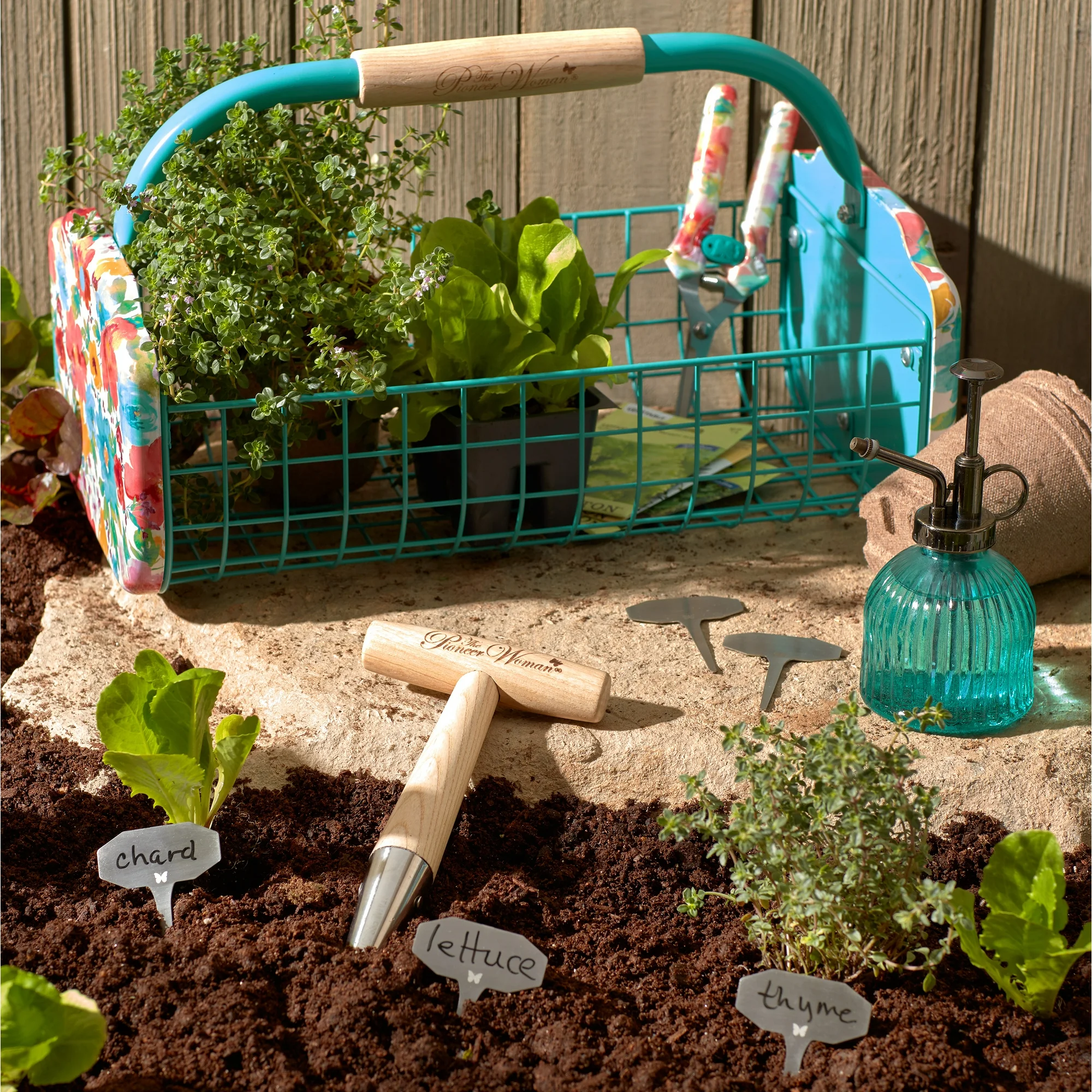 Gardening tools, plant markers labeled \“chard,\“ \“lettuce,\“ and \“thyme,\“ and a vibrant teal garden basket filled with plants are arranged on soil.