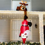 An inflatable Santa Claus hanging upside down from the roof, held by a reindeer, surrounded by white string lights and garland.