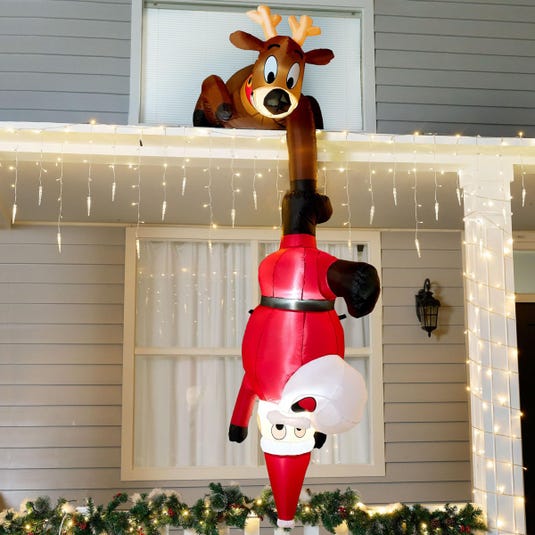 An inflatable Santa Claus hanging upside down from the roof, held by a reindeer, surrounded by white string lights and garland.