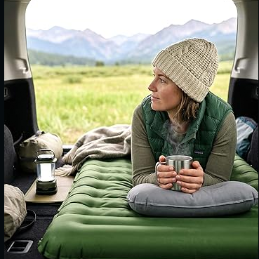 A person relaxes on an inflatable green mattress in the back of a vehicle, holding a mug. Nearby are a lantern, a pillow, and a backpack, with a scenic mountain view in the background.