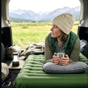 A person relaxes on an inflatable green mattress in the back of a vehicle, holding a mug. Nearby are a lantern, a pillow, and a backpack, with a scenic mountain view in the background.