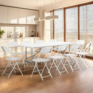 A modern kitchen features a long white folding table surrounded by eight white folding chairs on hardwood flooring, next to large windows with blinds.