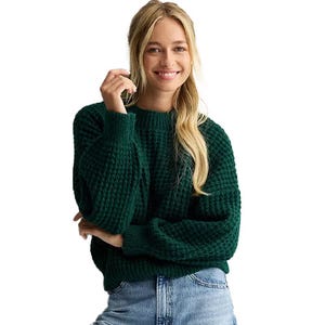 A woman is wearing a textured green sweater and light blue jeans, smiling with one hand lifted.