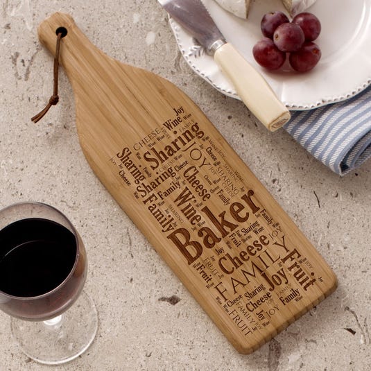 Custom wine cutting board shaped like a wine bottle with engraved words like \“Baker,\“ \“Sharing,\“ \“Cheese,\“ \“Joy,\“ \“Family,\“ and \“Wine.\“ Accompanied by a plate with cheese, grapes, and a glass of red wine.