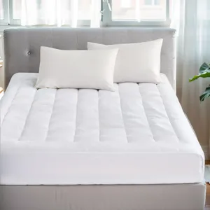A white quilted mattress topper with two matching pillows is on a light gray upholstered bed frame, set in a bright room with wooden floors and a potted plant nearby.