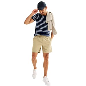 A man wears a navy striped t-shirt, khaki shorts, white sneakers, and a baseball cap, with a sweater draped over his shoulder.