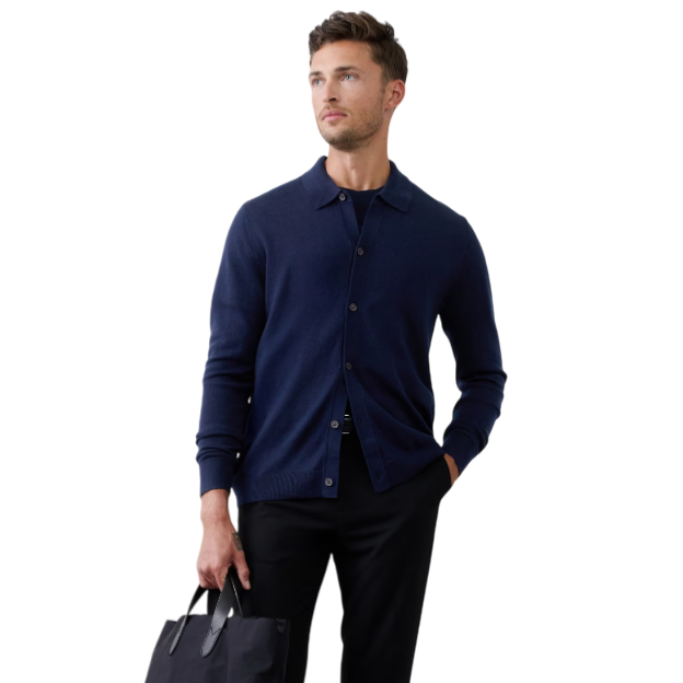 A man is wearing a navy button-up cardigan over a dark shirt with black trousers and black loafers, holding a black tote bag.