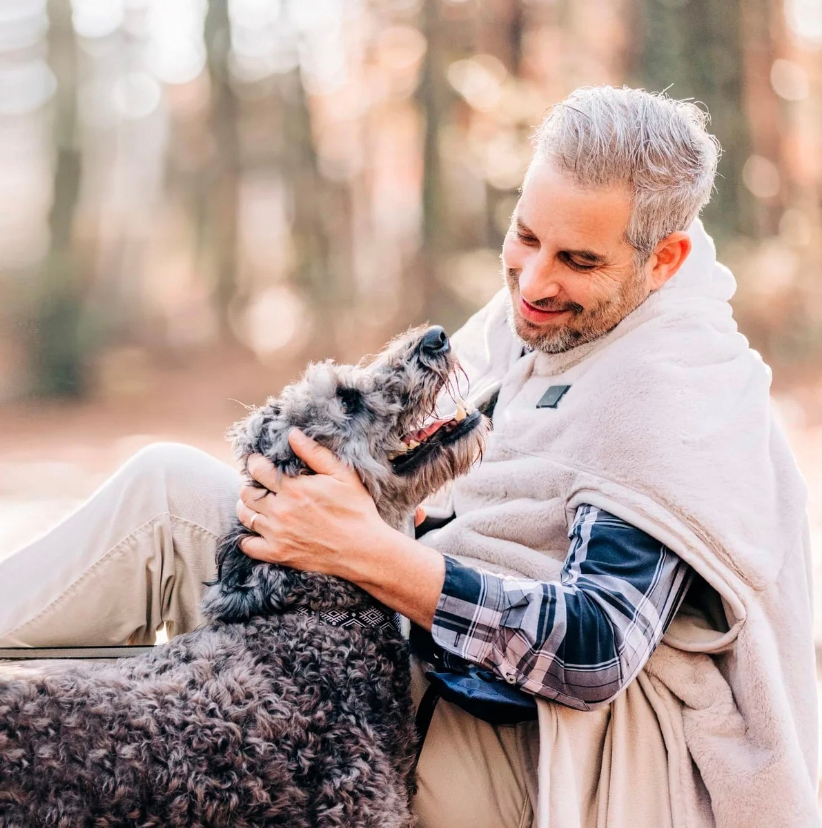 A man embracing a dog outdoors, both appear happy. The man is wearing a beige fleece jacket and a plaid shirt.