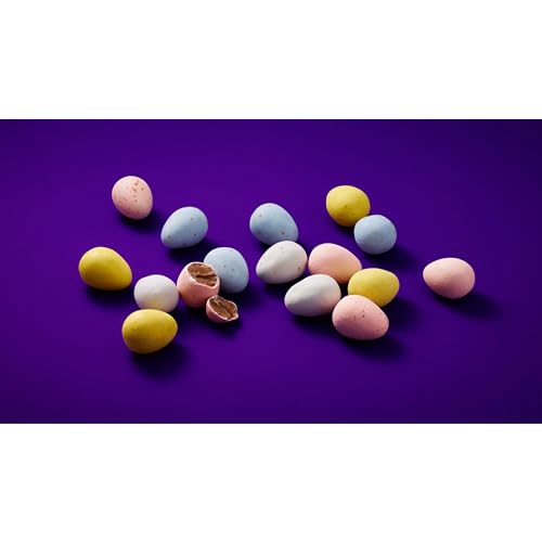 Assorted pastel-colored candy-coated chocolate eggs are scattered on a purple background, with one egg broken open to reveal chocolate inside.