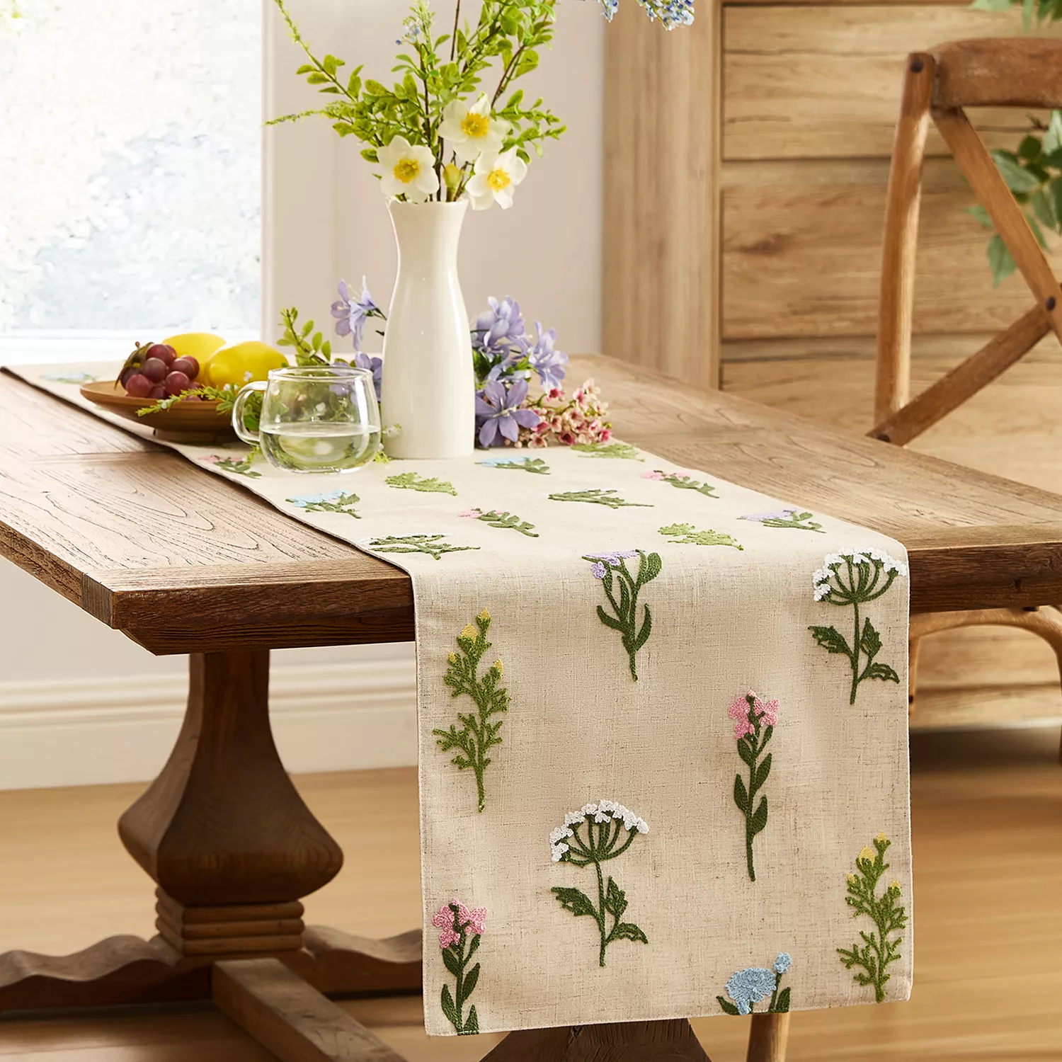 A wooden dining table with a floral-embroidered table runner holds a white vase with flowers, a bowl of fruit, and a glass mug of water.