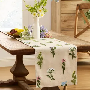 A wooden dining table with a floral-embroidered table runner holds a white vase with flowers, a bowl of fruit, and a glass mug of water.