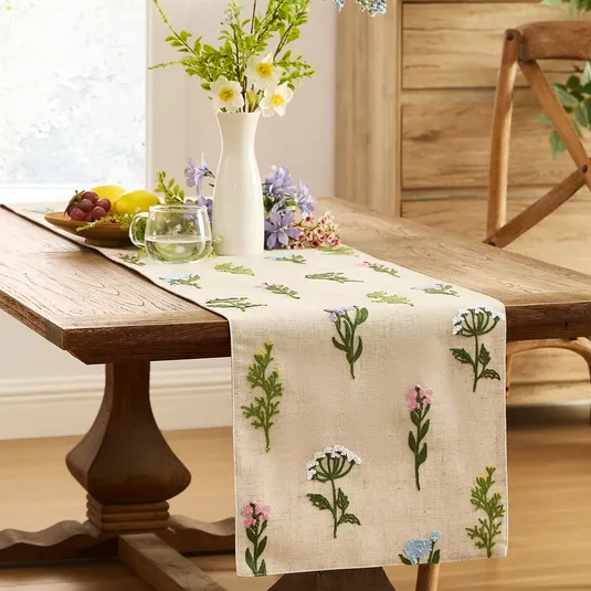 A wooden dining table with a floral-embroidered table runner holds a white vase with flowers, a bowl of fruit, and a glass mug of water.