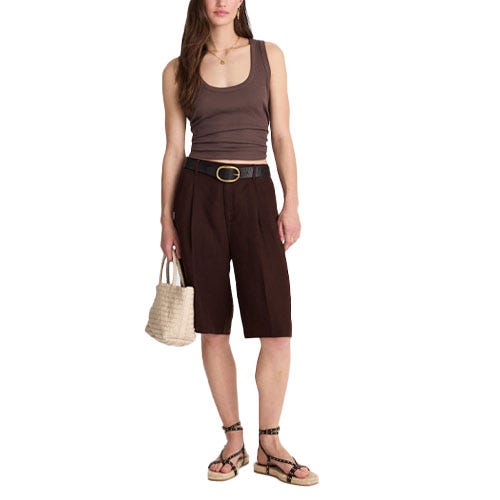 A woman is wearing a brown tank top, dark knee-length shorts with a black belt, and strappy flat sandals. She's holding a woven handbag.