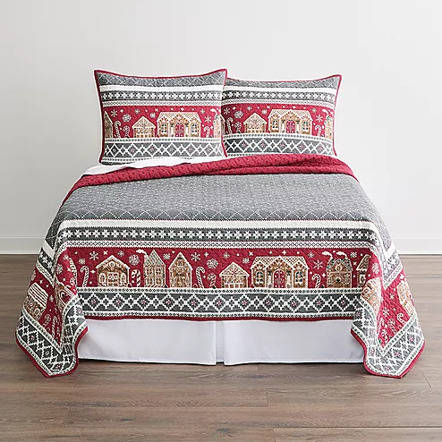 A bed is covered with a quilt featuring a festive red and grey pattern with snowflakes and village houses, accompanied by matching pillow shams.