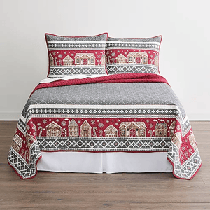 A bed is covered with a quilt featuring a festive red and grey pattern with snowflakes and village houses, accompanied by matching pillow shams.