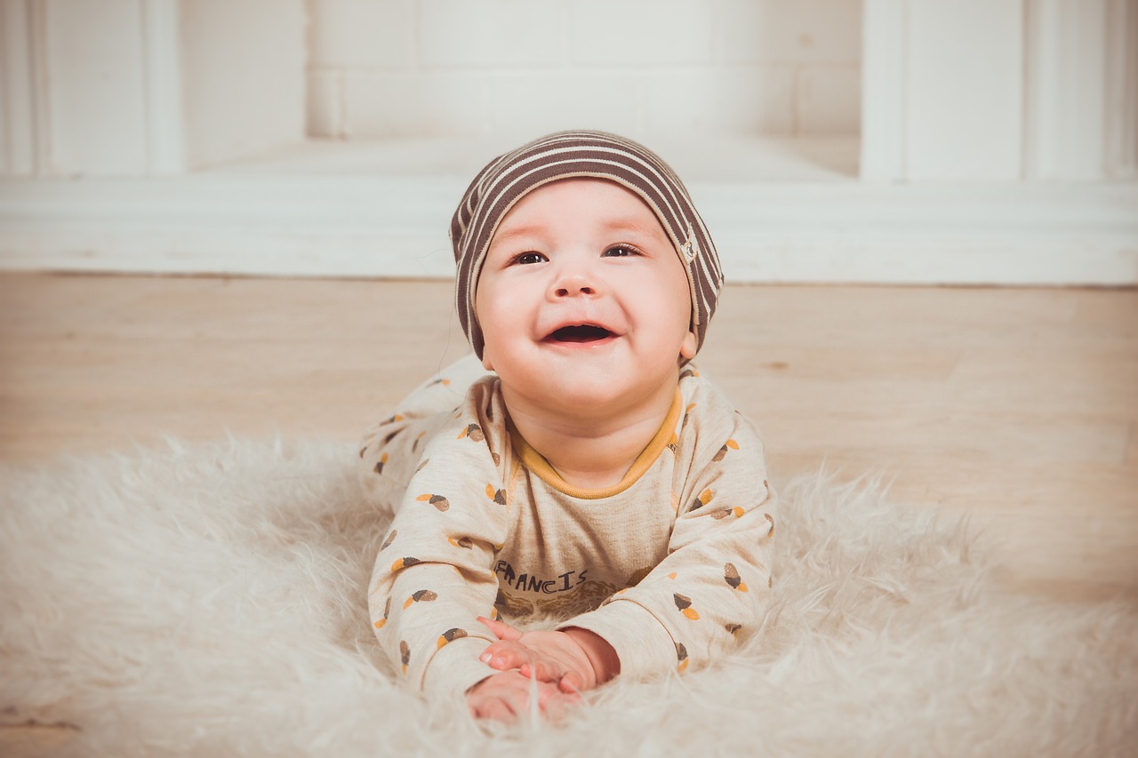 A smiling baby in a striped hat and a beige outfit with leaf patterns, lying on a white fluffy rug.