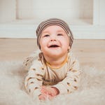 A smiling baby in a striped hat and a beige outfit with leaf patterns, lying on a white fluffy rug.