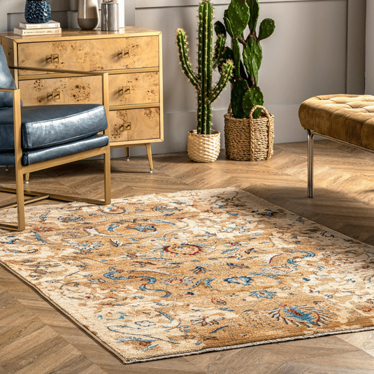 A patterned area rug with a floral design is on the floor, accompanied by a beige wooden dresser, a blue chair with gold frame, a sleek bench, and a tall cactus in a woven basket.