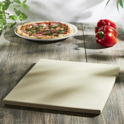 A pizza on a plate with basil leaves on a wooden surface, beside a red bell pepper and a square pizza stone.