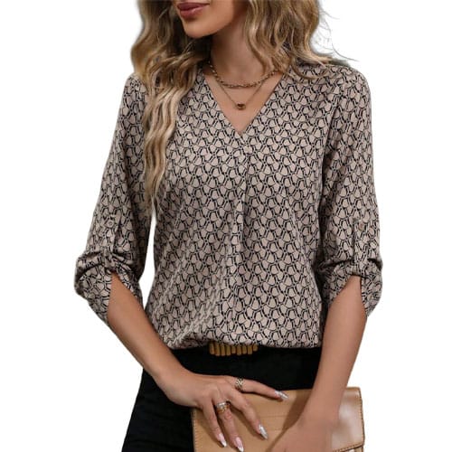 A woman is wearing a patterned, long-sleeved blouse with a V-neck and rolled-up sleeves, holding a tan clutch. She also wears minimal jewelry, including rings and layered necklaces.