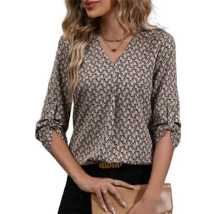 A woman is wearing a patterned, long-sleeved blouse with a V-neck and rolled-up sleeves, holding a tan clutch. She also wears minimal jewelry, including rings and layered necklaces.